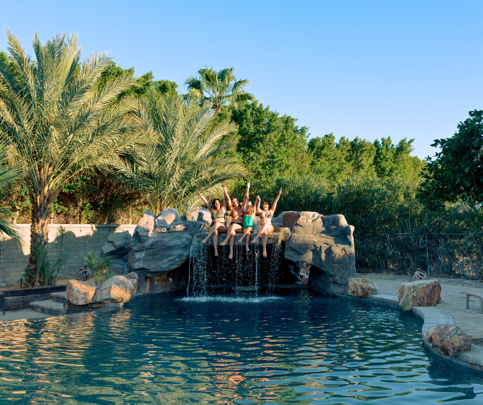 Group of women sitting on top of a pool waterfall feature, raising their arms in the air